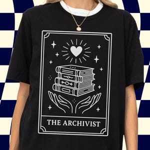 May include: Black t-shirt featuring a white graphic of a stack of books, a heart, and hands, with stars and the text "THE ARCHIVIST" in a rectangular frame. The shirt has a white collar.