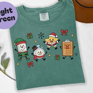 May include: A light green t-shirt featuring a graphic print of four cartoon characters: an elf-dressed book, a snowman with a stopwatch face, a star in a Santa suit, and a gingerbread clipboard. The shirt has a Comfort Colors label at the neck.