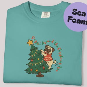 May include: Seafoam green t-shirt with a graphic of a pug dog decorating a Christmas tree. The pug is wearing a red sweater. The text "Puggin' Around the Christmas Tree" is in a circular design. A purple circle with the words "Sea Foam" is in the upper right.