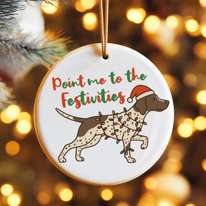 Könnte beinhalten: Weißer Keramik-Weihnachtsschmuck mit einem braun-weißen Hund, der einen Weihnachtsmannhut und Lichterketten trägt. Der Text "Point me to the Festivities" ist in Rot und Grün gehalten.