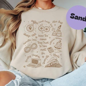 Peut inclure: Sweat-shirt couleur sable avec un motif graphique comprenant des illustrations de satellite, symbole infini, ordinateur, train et autres symboles scientifiques. Le texte comprend des noms comme Gladys West et Katherine Johnson, et la phrase "know the names they tried to take credit for."