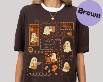 Puede incluir: Camiseta marrón con cuello blanco con un diseño de fantasmas de dibujos animados en marcos. El diseño incluye calabazas, hojas y el texto "Making Spirits Bright". Un círculo morado con la palabra "Brown" está en la parte superior derecha.