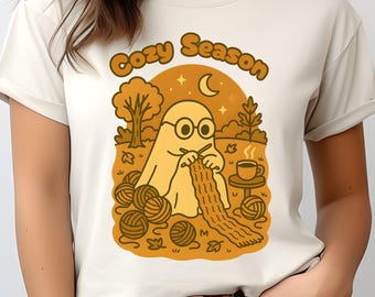 Può includere: T-shirt color crema con una grafica raffigurante un fantasma che lavora a maglia. Il fantasma indossa occhiali e lavora a maglia una sciarpa, circondato da gomitoli di lana, una tazza di caffè e le parole "Cozy Season".
