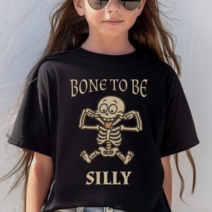 May include: Black t-shirt with the words "BONE TO BE SILLY" and a cartoon skeleton graphic. The skeleton is making a silly face with its tongue out. The shirt is a crew neck.