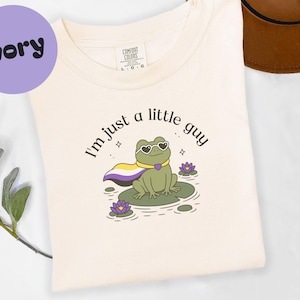 Puede incluir: Camiseta de color marfil con un gráfico de una rana con gafas de sol y una capa, sentada en una hoja de nenúfar. El texto "I'm just a little guy" está encima de la rana. Un círculo morado con la palabra "ivory" está en la parte superior izquierda.