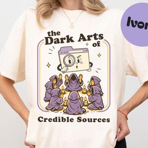 Könnte beinhalten: Elfenbeinfarbenes T-Shirt mit Cartoon-Grafik. Das Design zeigt eine Kamera mit Lupe, Figuren in lila Roben und den Text "the Dark Arts of Credible Sources". Ein lila Kreis sagt "ivory".