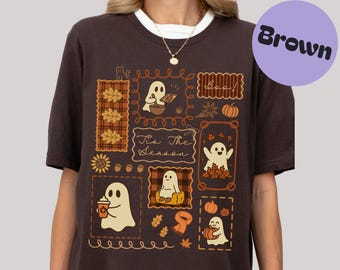 Könnte beinhalten: Braunes T-Shirt mit einem verspielten Design aus Cartoon-Geistern, Kürbissen und Herbstblättern. Das Design enthält den Text "Boo" und "Tis The Season". Das Shirt hat einen Rundhalsausschnitt und kurze Ärmel.
