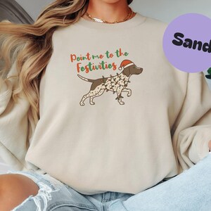 May include: Sand-colored sweatshirt featuring a cartoon dog wearing a Santa hat and Christmas lights. The text "Point me to the Festivities" is above the dog. A purple circle with the word "Sand" is in the upper right corner.