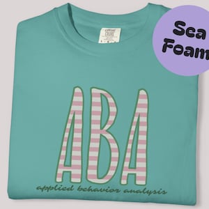 Könnte beinhalten: Meergrünes T-Shirt mit den Buchstaben "ABA" in einem rosa-weiß gestreiften Muster. Die Worte "applied behavior analysis" sind unter den Buchstaben aufgedruckt. Ein lila Kreis mit den Worten "Sea Foam" befindet sich in der oberen rechten Ecke.