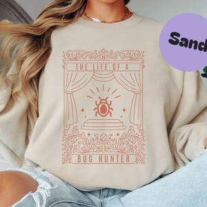 Puede incluir: Sudadera color arena con un gráfico rojo que dice "THE LIFE OF A BUG HUNTER". El gráfico presenta un escenario con una ilustración de un insecto. Un círculo morado con la palabra "Sand" está en la esquina superior derecha.