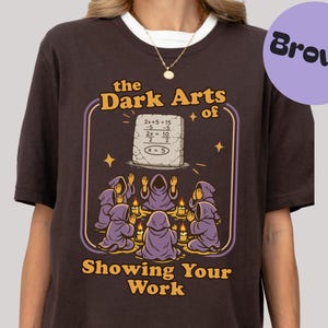 Peut inclure: T-shirt marron avec un motif représentant "the Dark Arts of Showing Your Work". Le design comprend une tablette de pierre avec une équation mathématique, des personnages en robes violettes, des bougies et des étoiles.