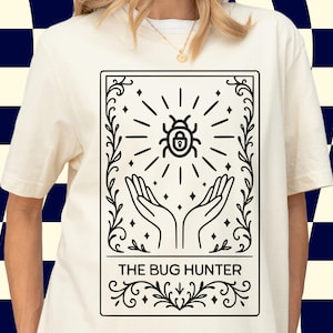 May include: A cream-coloured t-shirt with a black graphic. The graphic depicts a beetle with radiating lines, a lock, and open hands. The text "THE BUG HUNTER" is below. The shirt has short sleeves and a crew neck.