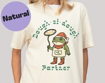 Op de afbeelding: Crèmekleurig T-shirt met een cartoon kikker karakter met een koksmuts, schort en laarzen. De kikker houdt een net met sterren vast en de tekst "Dough-si-dough Partner" staat op het shirt.