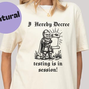 May include: Cream-colored t-shirt with the text "I Hereby Decree" above a cartoon knight holding a scroll. Below the knight, the text reads "testing is in session!" A purple circle with the word "Natural" is in the upper left corner.