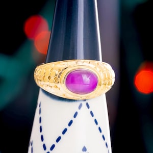 Puede incluir: Un anillo de oro con una banda texturizada y una gran piedra preciosa ovalada de color púrpura.