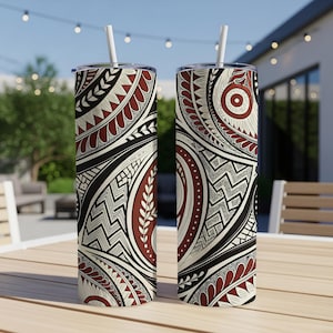 May include: Two tall, insulated tumblers with intricate tribal patterns in black, cream, and burgundy. Each tumbler has a white straw. The design features geometric shapes, lines, and leaf-like elements. The tumblers are on a wooden table outdoors.