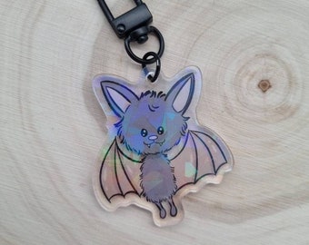 Könnte beinhalten: Ein holografischer Schlüsselanhänger mit einer Cartoon-Fledermaus mit einem süßen Ausdruck. Die Fledermaus ist grau mit schwarzen Augen und einer rosa Nase. Der Schlüsselanhänger hat einen schwarzen Metallverschluss.