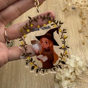 Könnte beinhalten: Ein klarer Acryl-Schlüsselanhänger mit einer Cartoon-Hexe. Die Hexe hat rotes Haar, einen schwarzen Hut und einen braunen Pullover, hält ein Buch und eine leuchtende Kaffeetasse. Der Text "Book Witch" steht darüber.