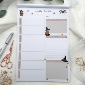 May include: A weekly planner notepad with a witchy theme. The notepad has sections for each day of the week, a "To Do" list, and a "Notes" section. Decorative elements include stars, a witch, and a witch's hat.