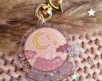 Könnte beinhalten: Ein transparenter Acryl-Schlüsselanhänger mit einem goldenen Metallverschluss. Der Schlüsselanhänger zeigt ein pink- und lilafarbenes Glitzerdesign mit einem Halbmond, Sternen und Wolken.
