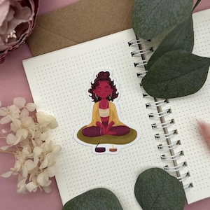 May include: A sticker featuring a cartoon illustration of a person meditating in a lotus position. The person has brown skin, long brown hair, and is wearing a yellow shirt and a red skirt. The sticker is on a white background with a dotted pattern.