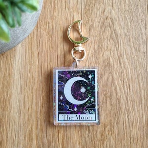 K&ouml;nnte beinhalten: Ein klarer Acryl-Schl&uuml;sselanh&auml;nger mit einem Tarotkarten-Design. Die Karte zeigt einen Halbmond, Sterne und Wolken auf einem lila und schwarzen Hintergrund. Der Text "The Moon" befindet sich unten, mit einem goldfarbenen Verschluss.