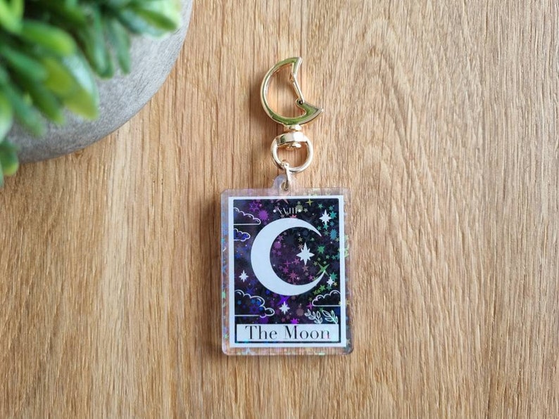 K&ouml;nnte beinhalten: Ein klarer Acryl-Schl&uuml;sselanh&auml;nger mit einem Tarotkarten-Design. Die Karte zeigt einen Halbmond, Sterne und Wolken auf einem lila und schwarzen Hintergrund. Der Text "The Moon" befindet sich unten, mit einem goldfarbenen Verschluss.