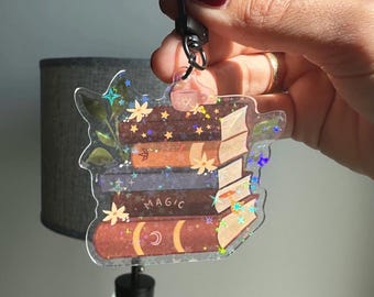 Könnte beinhalten: Transparenter Acryl-Schlüsselanhänger mit einem Stapel Bücher mit floralen und sternförmigen Akzenten. Die Bücher sind in verschiedenen Braun- und Lilatönen gehalten, mit dem Wort "MAGIC" sichtbar. Der Schlüsselanhänger hat einen schwarzen Verschluss.