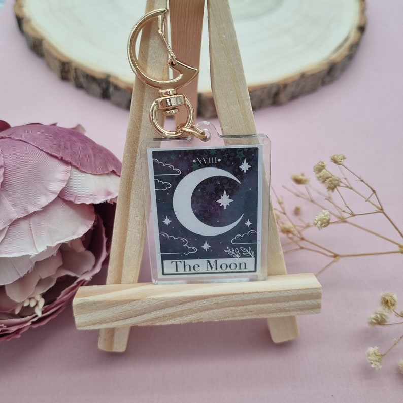 K&ouml;nnte beinhalten: Ein klarer Acryl-Schl&uuml;sselanh&auml;nger mit einem Tarotkarten-Design des Mondes. Die Karte hat einen Halbmond und Stern vor einem dunkelvioletten Hintergrund. Der Schl&uuml;sselanh&auml;nger hat einen goldfarbenen Verschluss.