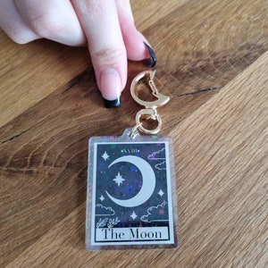 K&ouml;nnte beinhalten: Ein rechteckiger Schl&uuml;sselanh&auml;nger mit einem Tarotkarten-Design. Die Karte zeigt einen Halbmond, Sterne und Wolken auf einem dunklen, glitzernden Hintergrund. Der Text "The Moon" steht unten. Der Schl&uuml;sselanh&auml;nger hat einen goldfarbenen Verschluss.