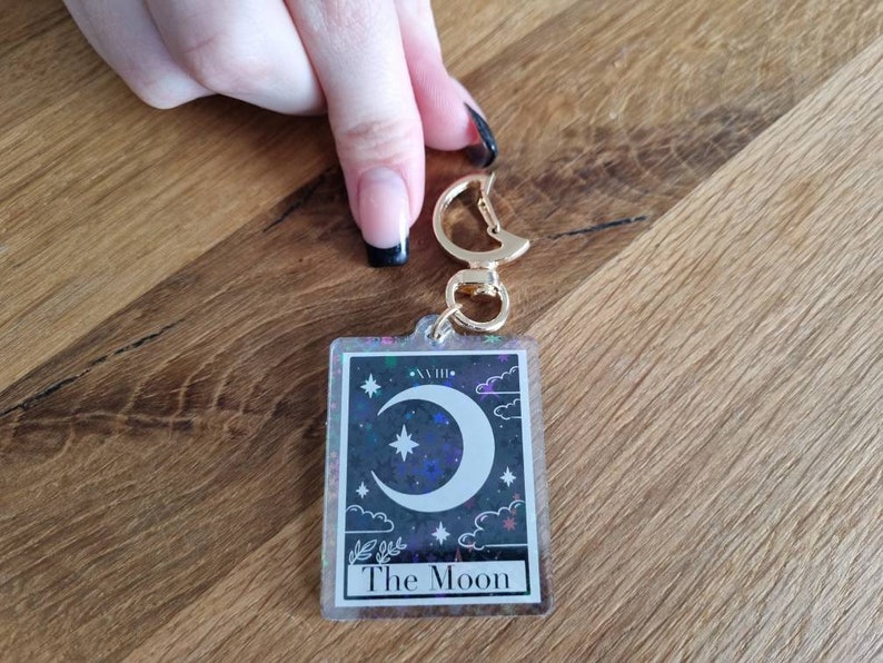 K&ouml;nnte beinhalten: Ein rechteckiger Schl&uuml;sselanh&auml;nger mit einem Tarotkarten-Design. Die Karte zeigt einen Halbmond, Sterne und Wolken auf einem dunklen, glitzernden Hintergrund. Der Text "The Moon" steht unten. Der Schl&uuml;sselanh&auml;nger hat einen goldfarbenen Verschluss.