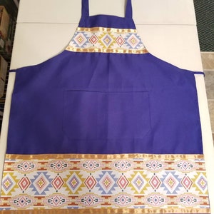 May include: A purple apron with a geometric pattern in shades of pink, blue, yellow, and brown. The apron has a large pocket on the front and a gold ribbon trim.