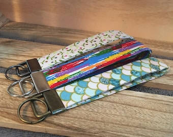 May include: Three fabric keychains with different patterns. The top keychain has a white background with small green and pink flowers. The middle keychain has a red, blue, yellow, and purple striped pattern. The bottom keychain has a white background with blue and gold scales.