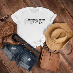 May include: White t-shirt with a black graphic of a Ford Bronco and the text "Bronco Hair Don't Care". The shirt is laid out on a wooden surface with a pair of brown cowboy boots, a brown cowboy hat, a pair of sunglasses, and a brown leather purse.