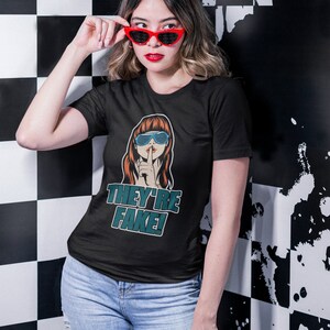 May include: Black t-shirt with a graphic of a woman wearing sunglasses and a finger over her lips, with the text "THEY'RE FAKE!". The person in the image is wearing red sunglasses and blue jeans.