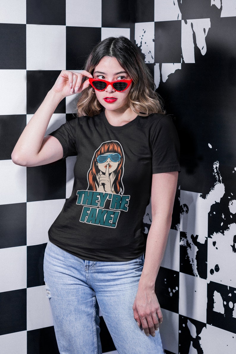May include: Black t-shirt with a graphic of a woman wearing sunglasses and a finger over her lips, with the text "THEY'RE FAKE!". The person in the image is wearing red sunglasses and blue jeans.