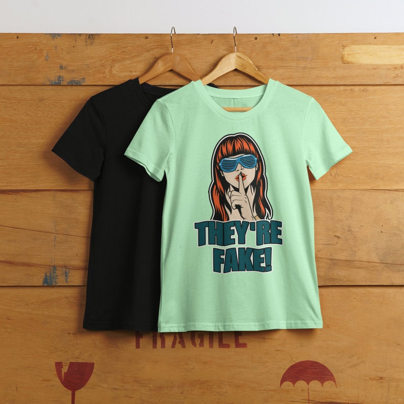May include: Two t-shirts hanging on wooden hangers. One is black, the other is light green with a graphic of a woman wearing blue sunglasses and the text "THEY'RE FAKE!" in teal. The background is a wooden wall.