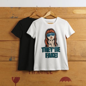May include: Two t-shirts hanging on wooden hangers. One is black, the other is white with a graphic of a woman with red hair and blue sunglasses, gesturing for silence. The text "THEY'RE FAKE!" is below the graphic.