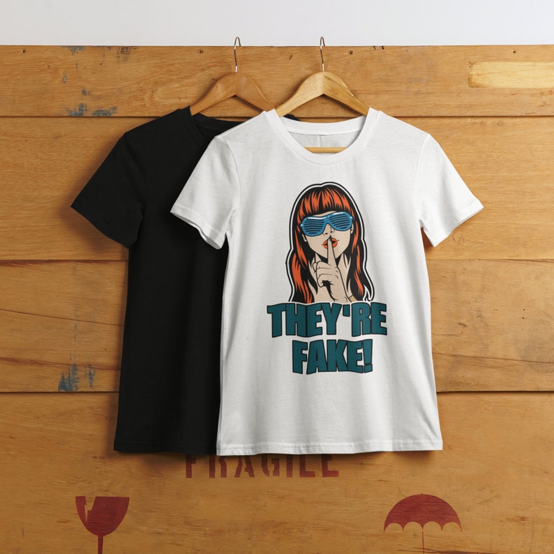 May include: Two t-shirts hanging on wooden hangers. One is black, the other is white with a graphic of a woman with red hair and blue sunglasses, gesturing for silence. The text "THEY'RE FAKE!" is below the graphic.