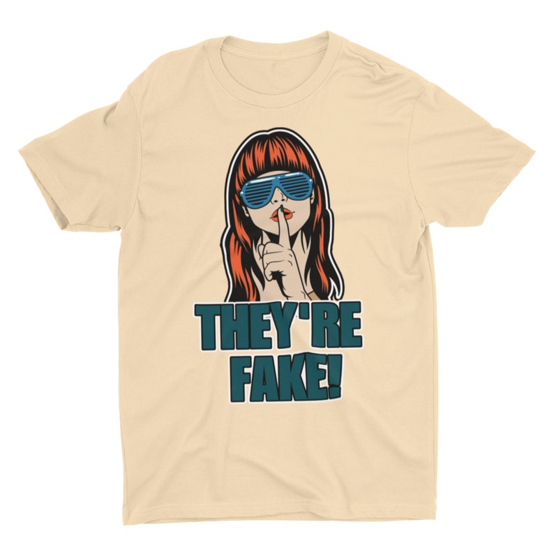 May include: A cream-colored t-shirt featuring a pop art style illustration. The graphic depicts a woman with red hair and blue sunglasses, finger to her lips, with the text "THEY'RE FAKE!" in teal.
