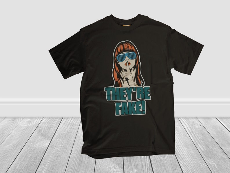 May include: Black t-shirt featuring a pop art style illustration of a woman with red hair and blue sunglasses, finger to her lips. The text "THEY'RE FAKE!" is printed below the image in teal and white.
