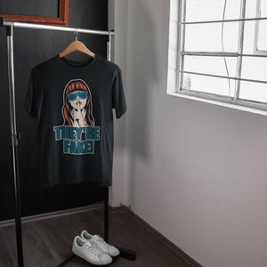 May include: Black t-shirt with a graphic of a woman wearing blue sunglasses and the text "THEY'RE FAKE!" displayed in teal. The shirt hangs on a wooden hanger on a clothing rack. White sneakers are placed on the floor.