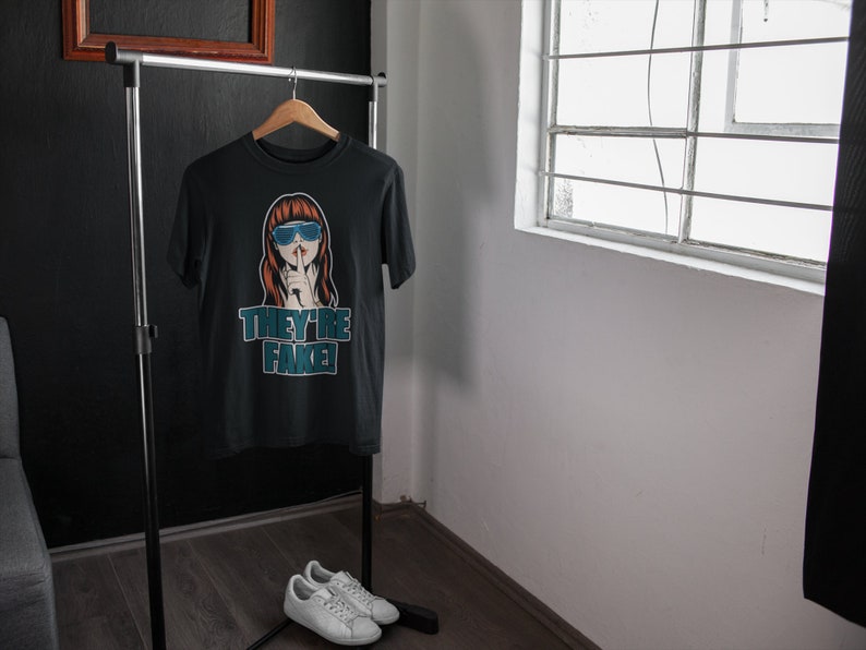 May include: Black t-shirt with a graphic of a woman wearing blue sunglasses and the text "THEY'RE FAKE!" displayed in teal. The shirt hangs on a wooden hanger on a clothing rack. White sneakers are placed on the floor.