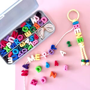 May include: A personalised keyring spelling "IRENE" in colourful letter beads. A matching beaded strand spells out "MERP" and "POP". A container of alphabet beads and numbers sits nearby on a pink surface.