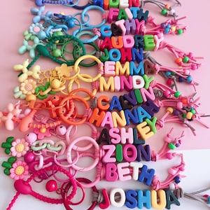 May include: A collection of colorful keychains with names spelled out in large, block letters. The keychains feature various designs, including flowers, balloons, and circles, in shades of pink, blue, green, yellow, and orange. The names include Aiden, Beth, and Joshua.