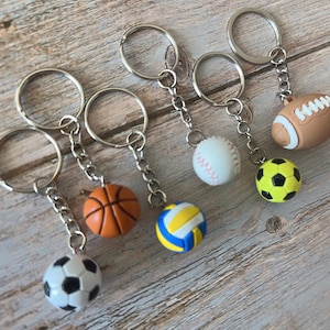Op de afbeelding: Een verzameling sleutelhangers met sportthema's, met miniatuur basketbal-, voetbal-, volleybal-, honkbal- en American football-ballen. Elke sleutelhanger heeft een zilverkleurige ring en ketting, en de sportballen zijn in hun respectievelijke kleuren.
