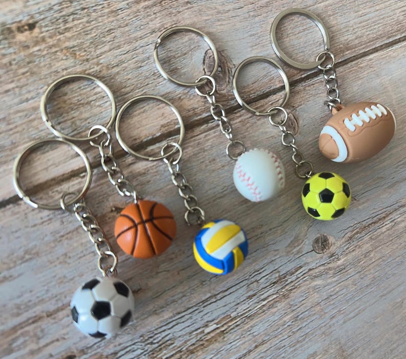 Op de afbeelding: Een verzameling sleutelhangers met sportthema's, met miniatuur basketbal-, voetbal-, volleybal-, honkbal- en American football-ballen. Elke sleutelhanger heeft een zilverkleurige ring en ketting, en de sportballen zijn in hun respectievelijke kleuren.
