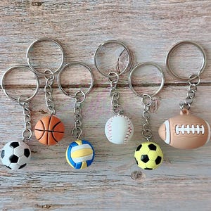 Op de afbeelding: Collectie sportthema sleutelhangers met een voetbal, basketbal, volleybal, honkbal, gele voetbal en American football. Elke sleutelhanger heeft een zilverkleurige ring en ketting.