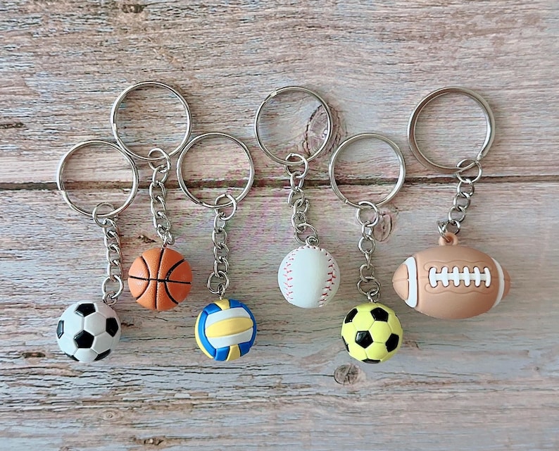 Op de afbeelding: Collectie sportthema sleutelhangers met een voetbal, basketbal, volleybal, honkbal, gele voetbal en American football. Elke sleutelhanger heeft een zilverkleurige ring en ketting.