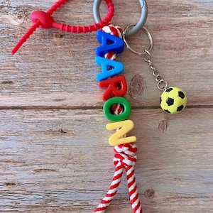 Op de afbeelding: Een gepersonaliseerde sleutelhanger met de naam "AARON" in kleurrijke letterkralen. De sleutelhanger bevat een voetbalbedel en een rood-wit gestreept touwontwerp. Een zilverkleurige sleutelring en een rode siliconen lus maken het ontwerp compleet.